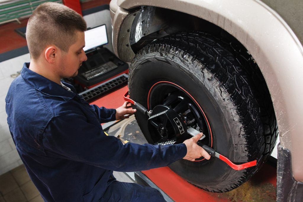 A mechanic in a blue uniform mounts a wheel alignment clamp and sensor target onto the large tire of a vehicle in a service bay, with a diagnostic computer monitor visible in the background.