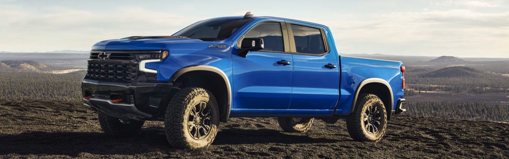 Blue Chevrolet Silverado 1500 pickup truck parked on rugged terrain overlooking a scenic mountain landscape.